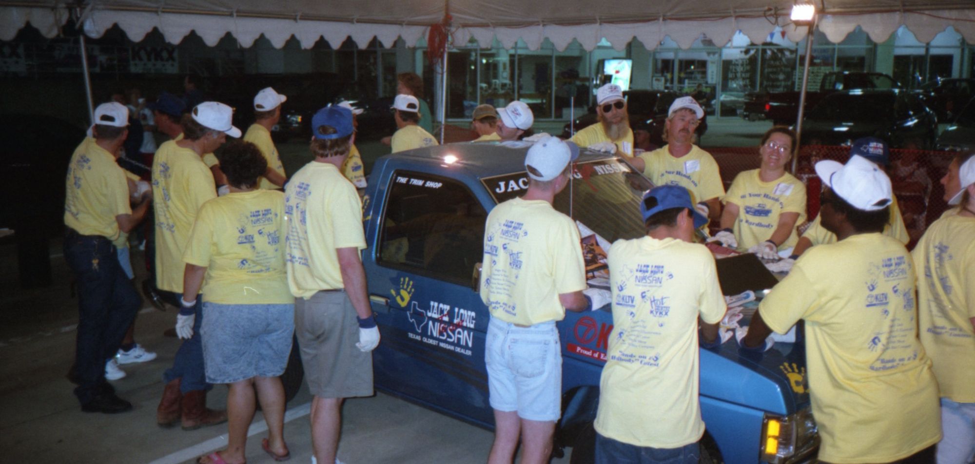 Contestants in yellow shirts around the blue pickup under a floodlight at night