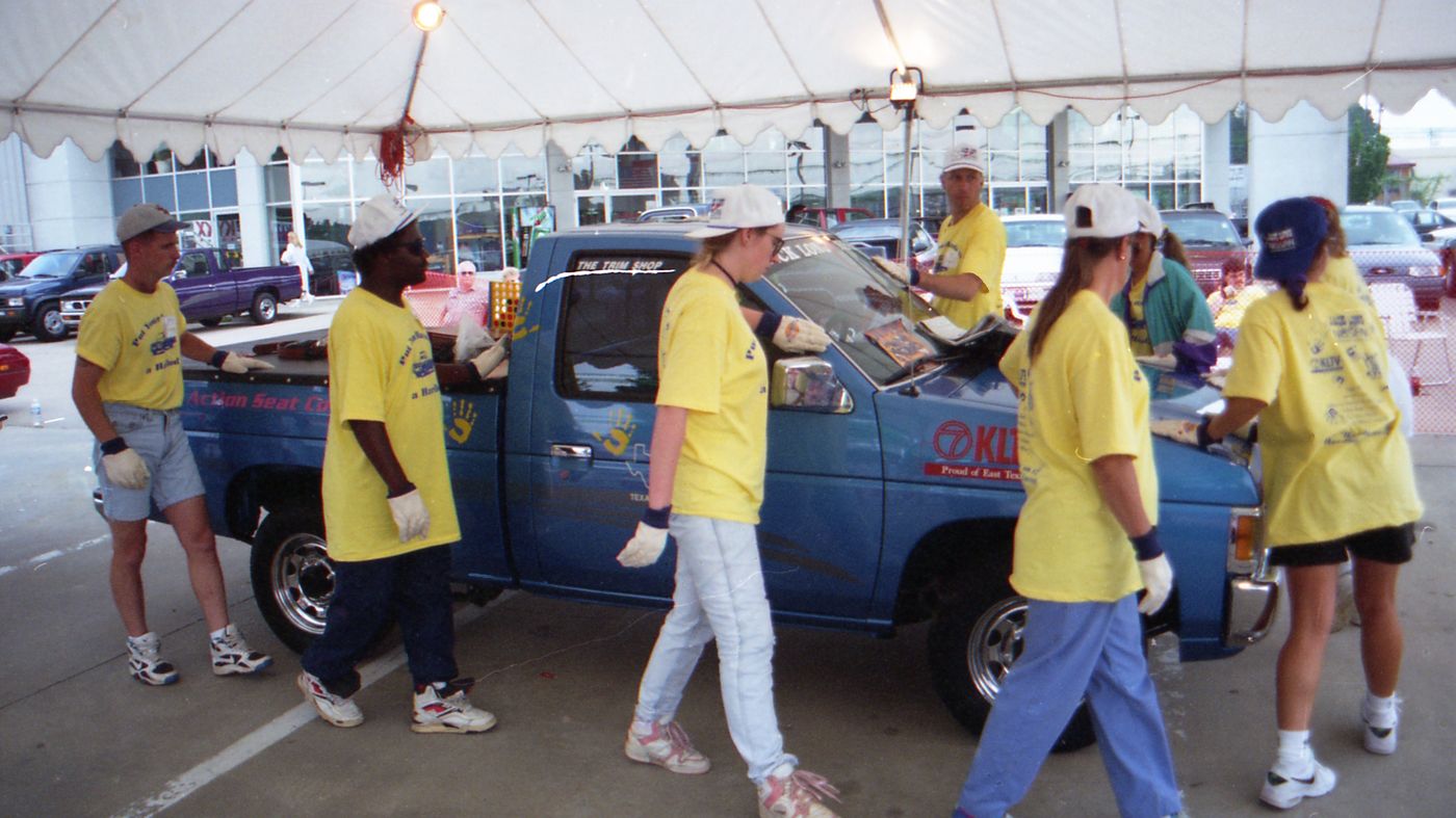 Contestants in yellow shirts walk around the blue pickup in daylight