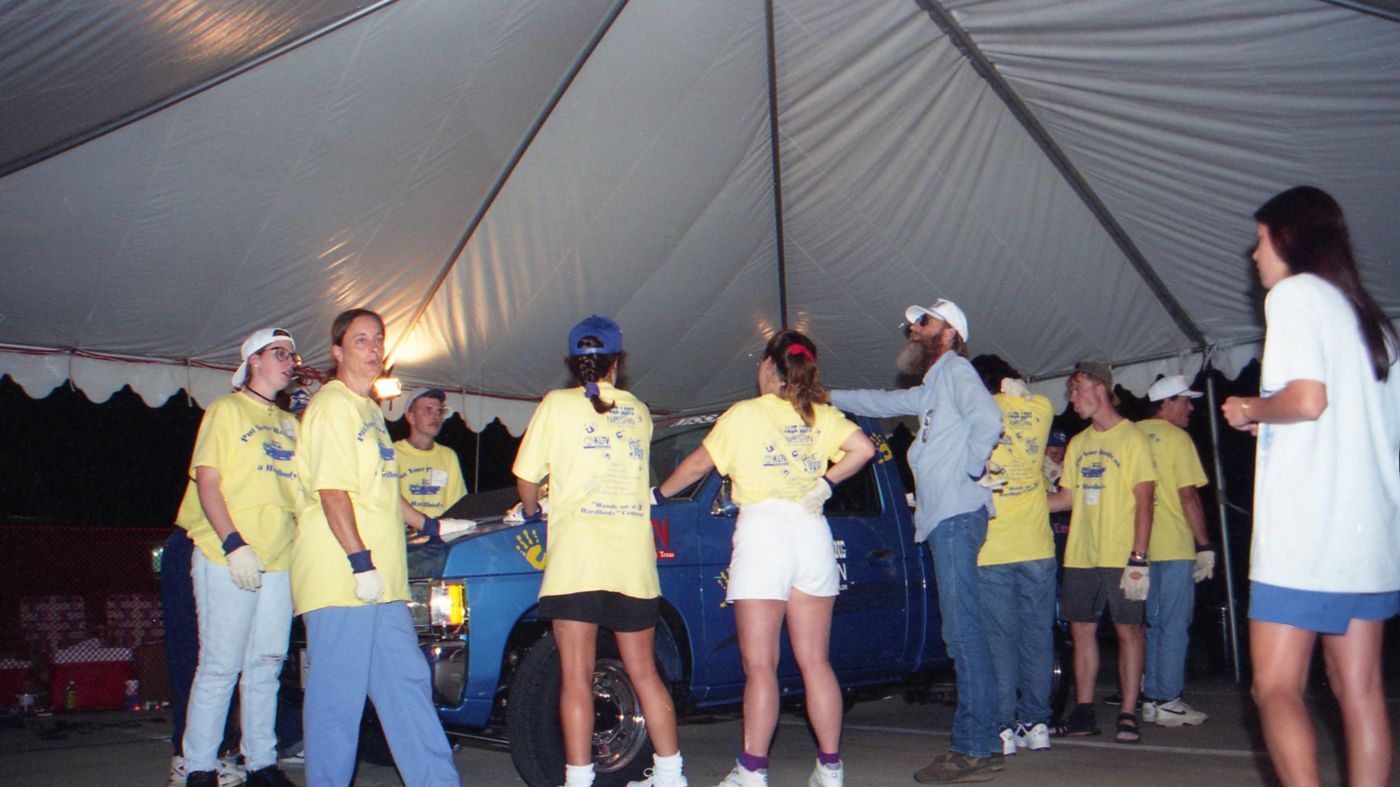 Contestants stand around the truck under the lit tent